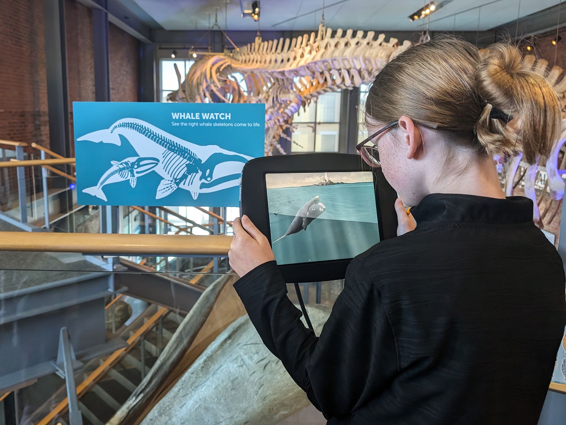 A teenage girl holds an iPad up to a sign and watches an augmented reality animation of a whale.