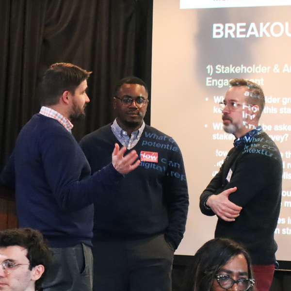 Three men talk in a circle in front of a projector screen that reads "breakout prompts"